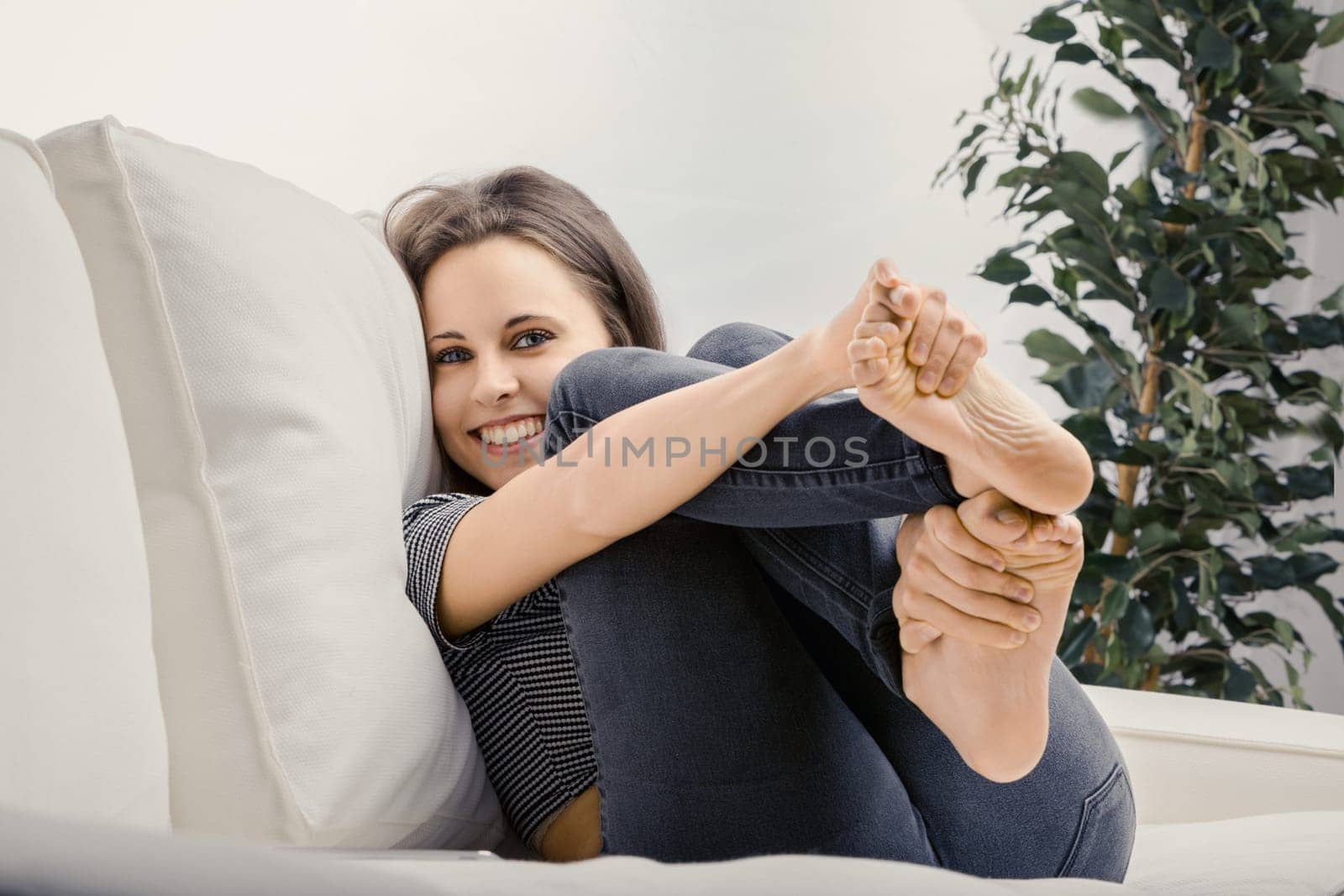Young woman sits happily on a white couch at home, smiling contentedly. Enjoying a peaceful day, she radiates relaxation and joy in her cozy living room