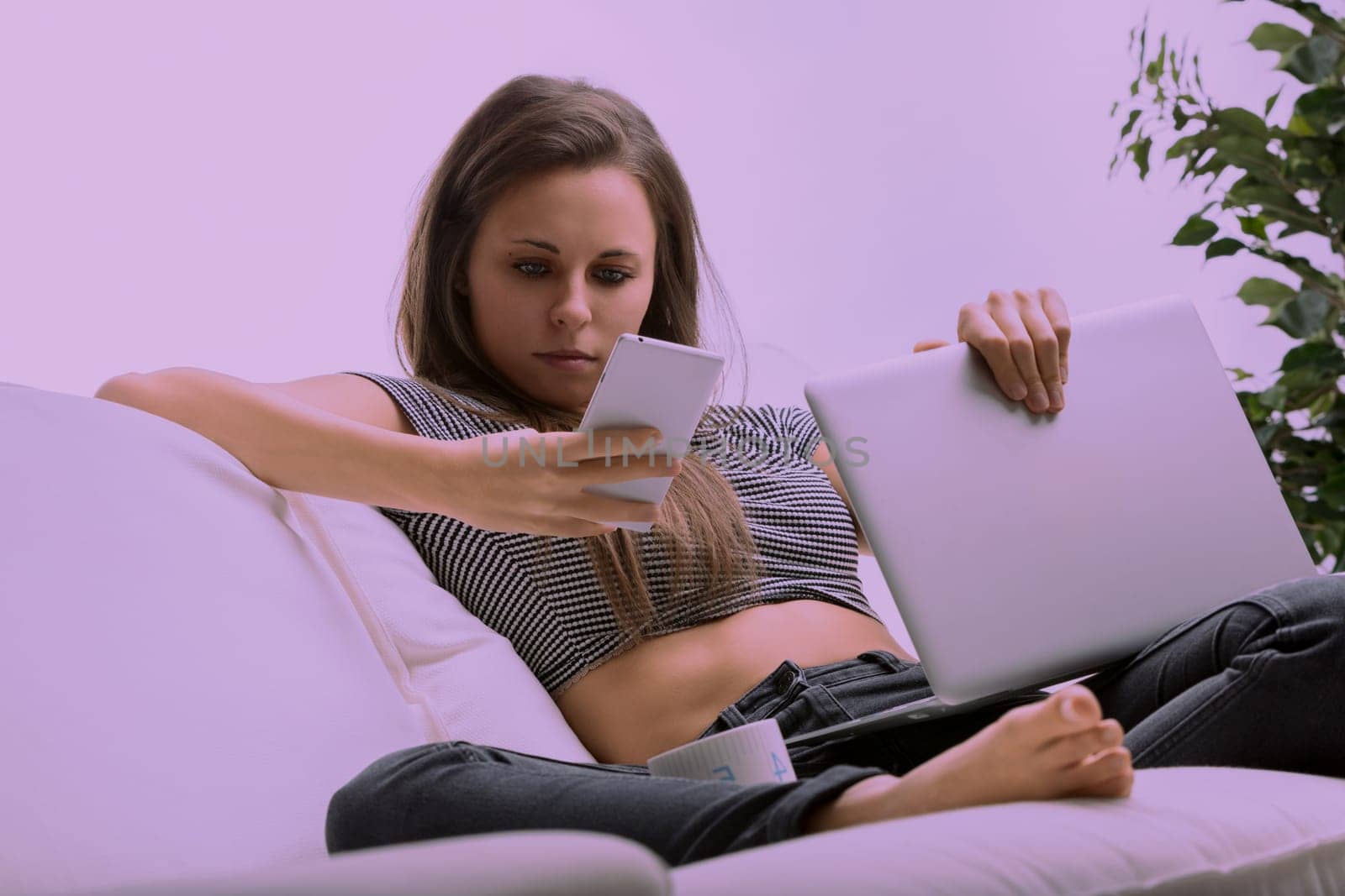 Young woman is lying on her couch, holding her laptop and browsing the internet on her smartphone. She has a neutral expression