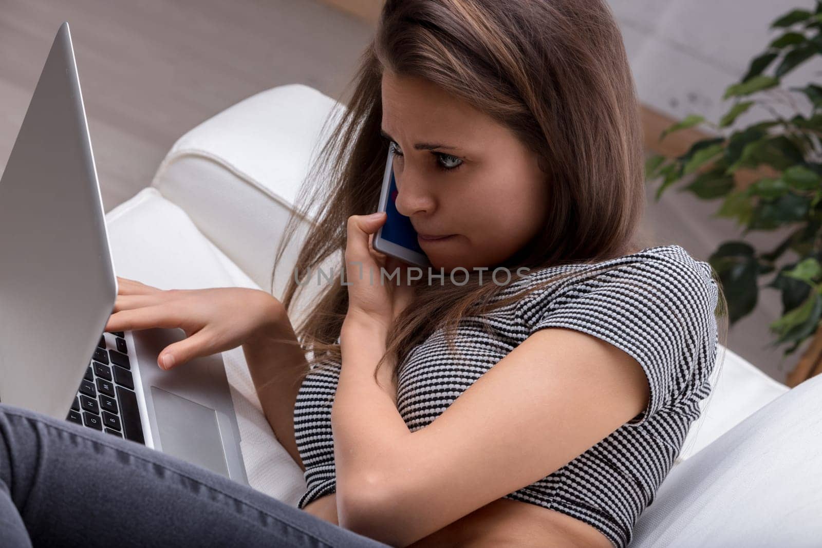 Stressed young woman multitasking on a sofa with laptop and phone, overwhelmed by remote work pressures
