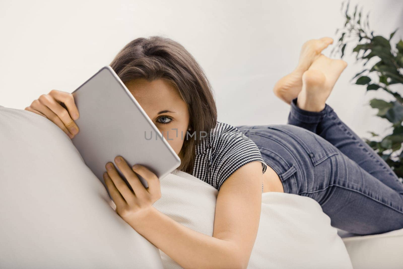 Young woman enjoying her digital tablet on a white couch at home, browsing, streaming, and connecting online in a casual and relaxed manner
