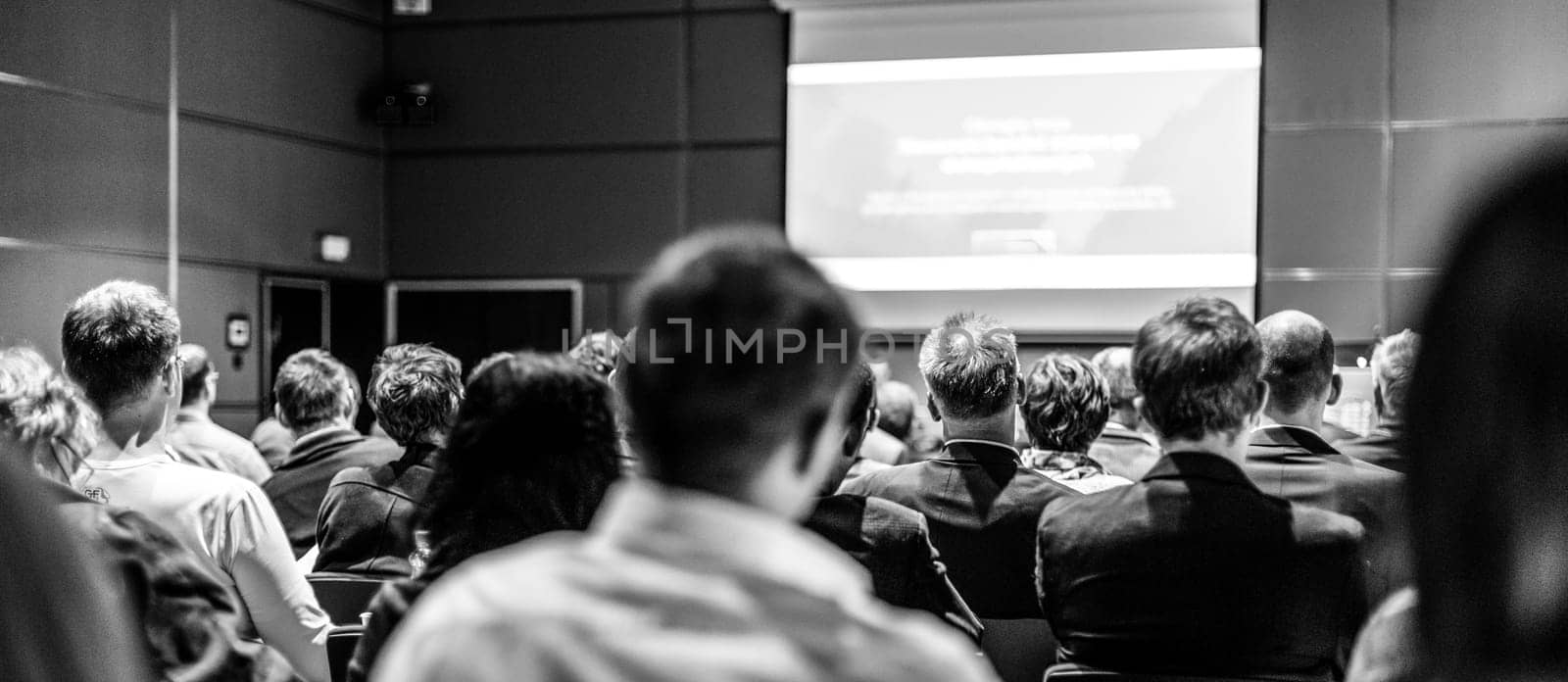 Business and entrepreneurship symposium. Speaker giving a talk at business meeting. Audience in conference hall. Rear view of unrecognized participant in audience.