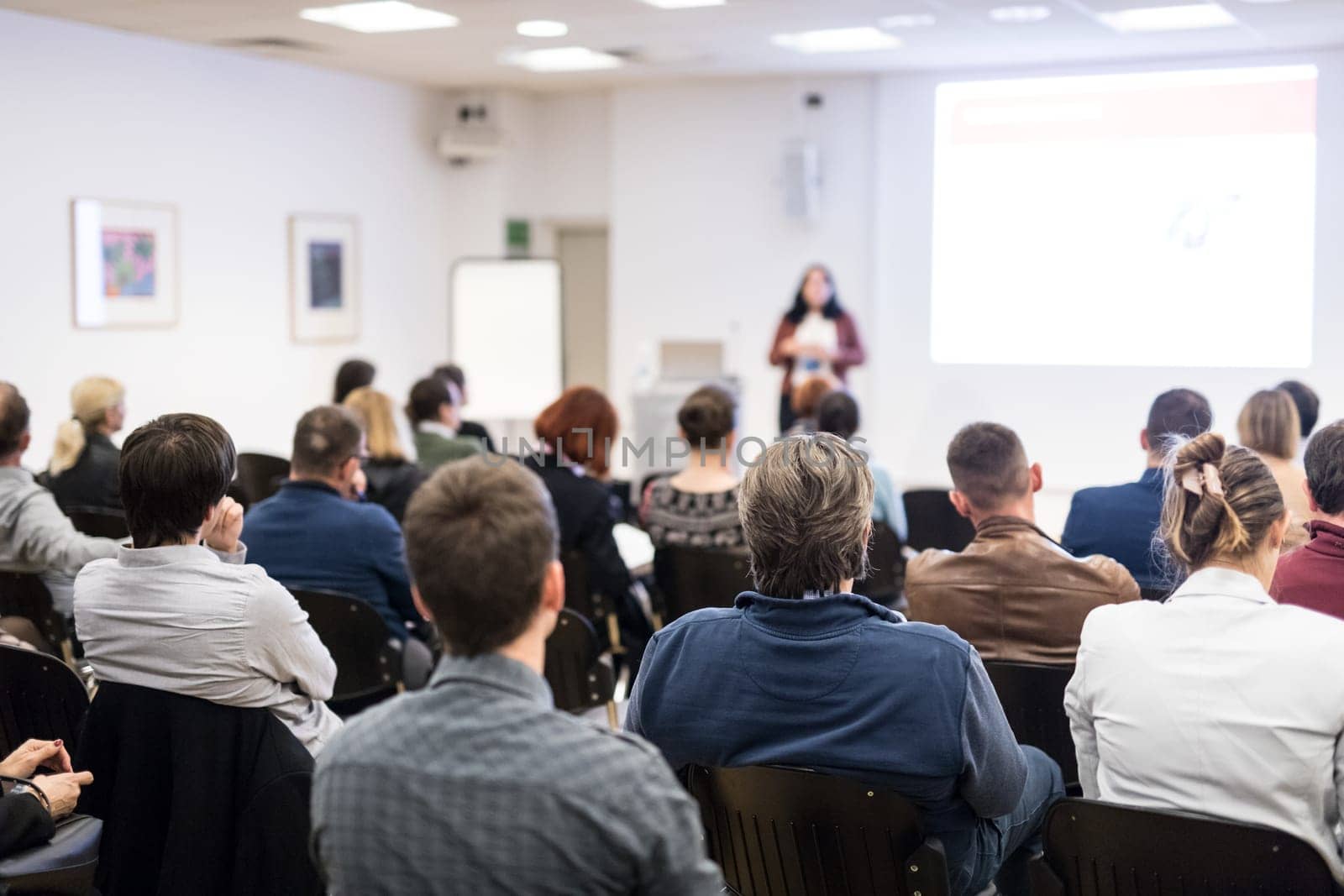 Business and entrepreneurship symposium. Female speaker giving a talk at business meeting. Audience in conference hall. Rear view of unrecognized participant in audience. Copy space on whitescreen.