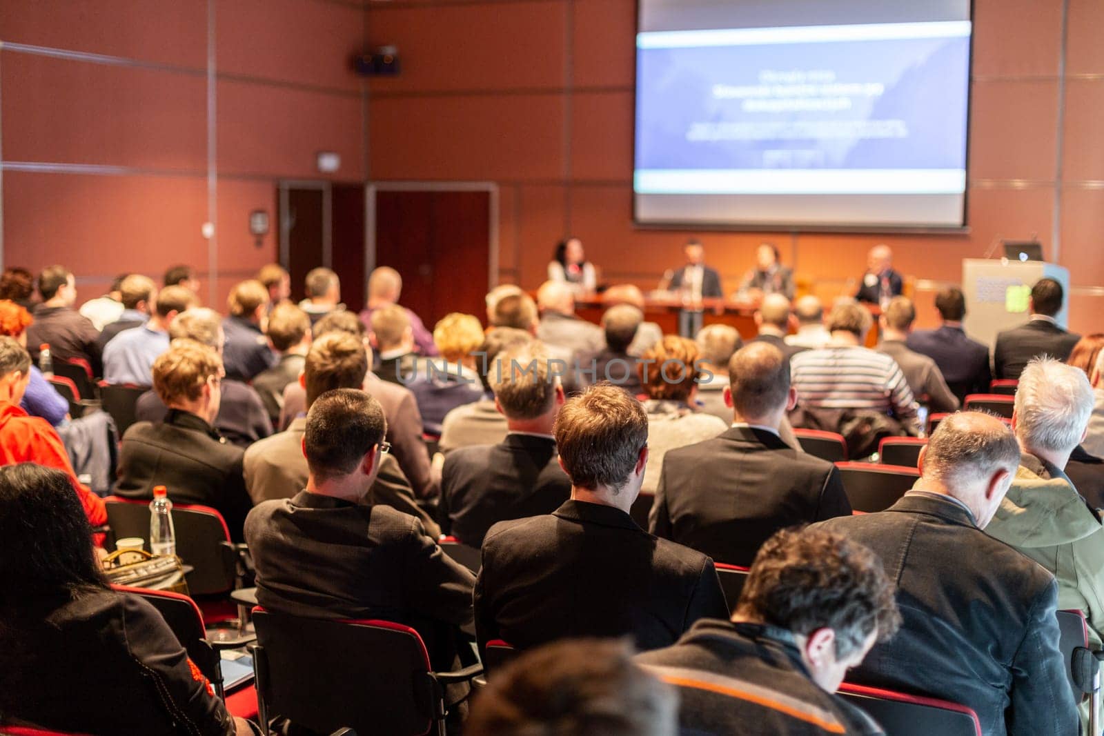 Business and entrepreneurship symposium. Speaker giving a talk at business meeting. Audience in the conference hall. Rear view of unrecognized participant in audience.