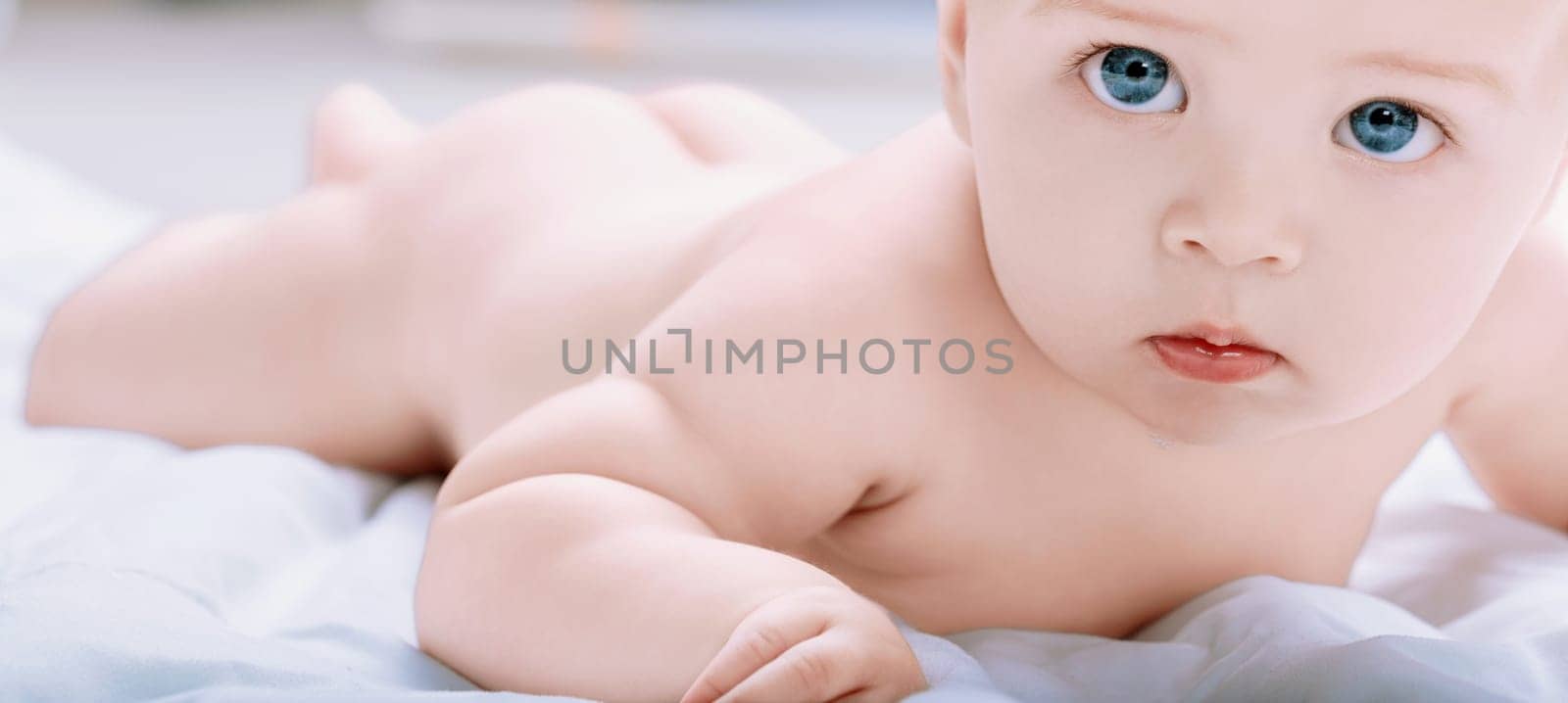 Adorable baby boy with bright blue eyes is lying on his stomach on a soft blanket, lifting his head and looking forward curiously