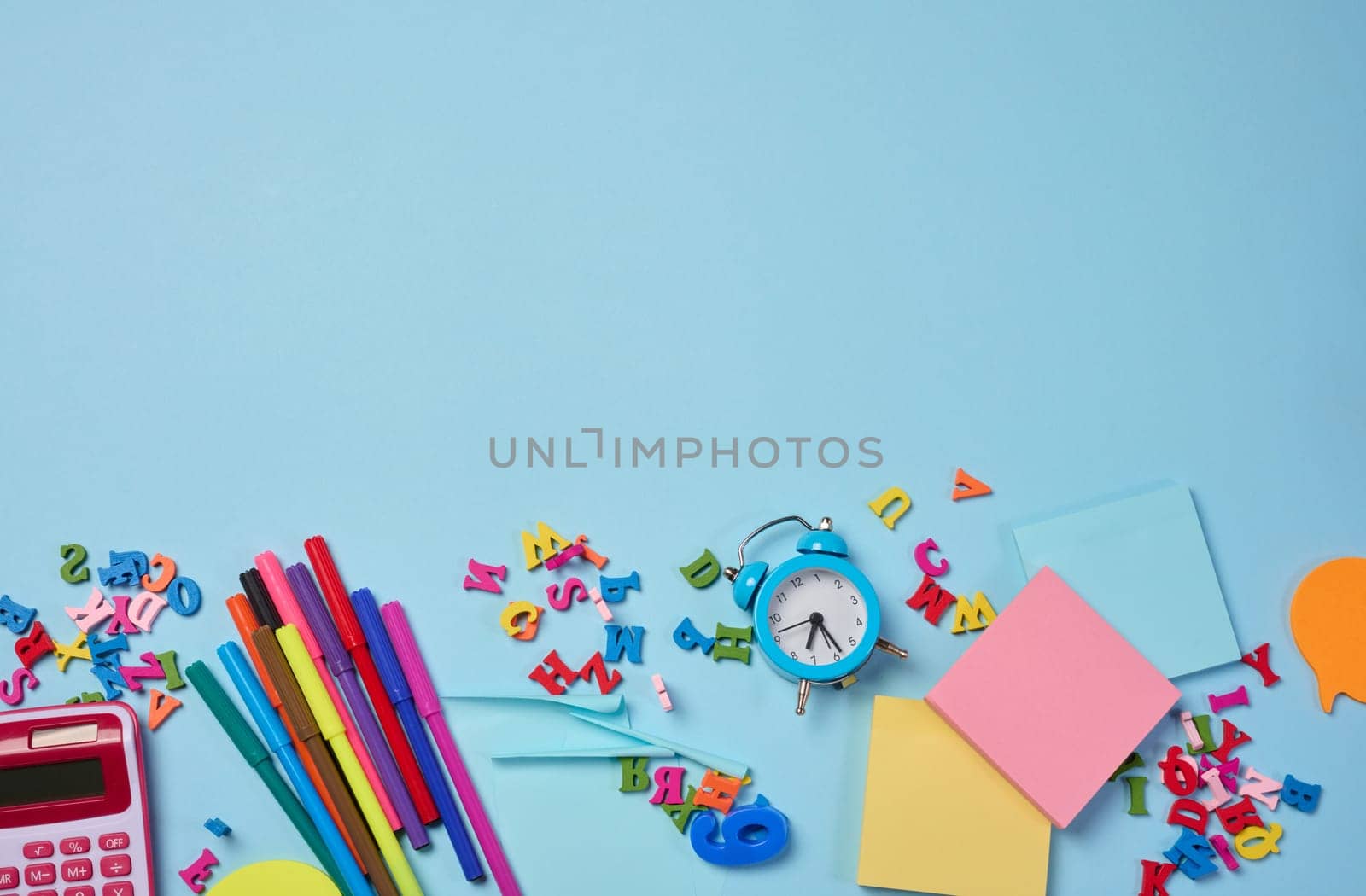 Stationery laid out on a blue background. Multi-colored felt-tip pens, calculator, alarm clock, sticky notes and wooden letters, preparing for study or work, organizing workspace