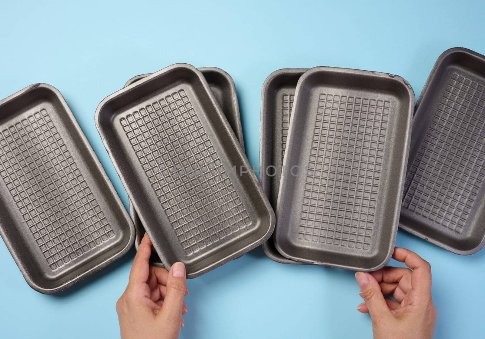 Hand holds black disposable polystyrene foam rectangular plate for packaging vegetables and meat on a blue background, top view