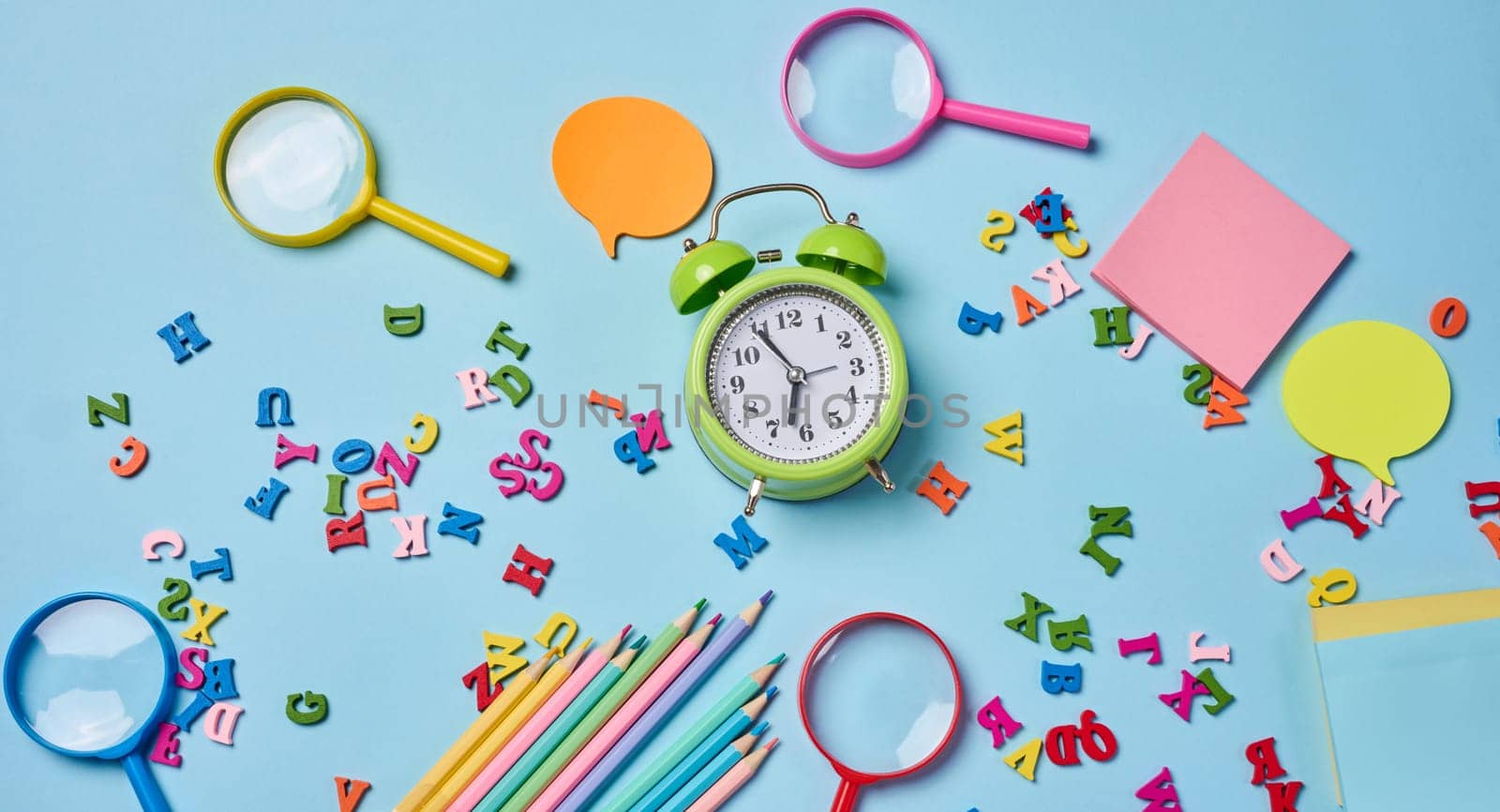 Stationery laid out on a blue background. Multi-colored felt-tip pens, alarm clock, sticky notes and wooden letters, preparing for study or work, organizing workspace