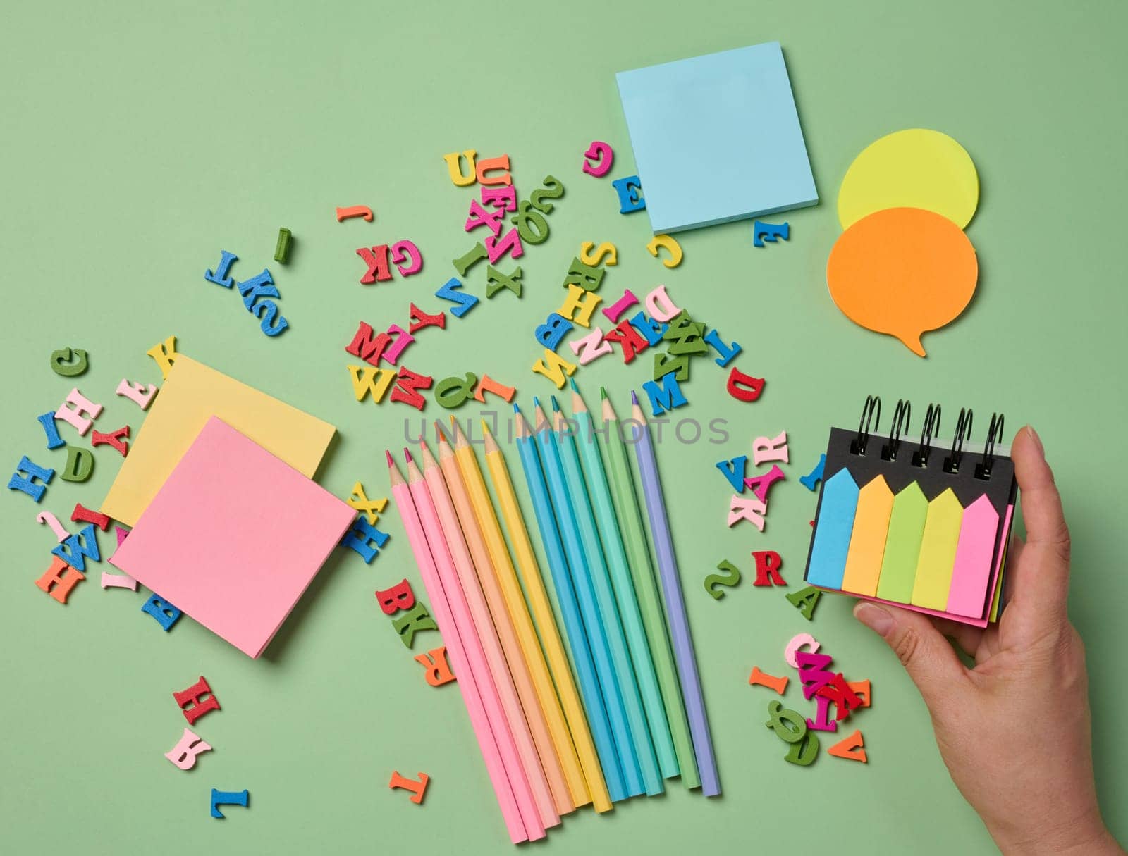 Stationery laid out on a green background. Multi-colored pencils ,  sticky notes and wooden letters, preparing for study or work, organizing workspace