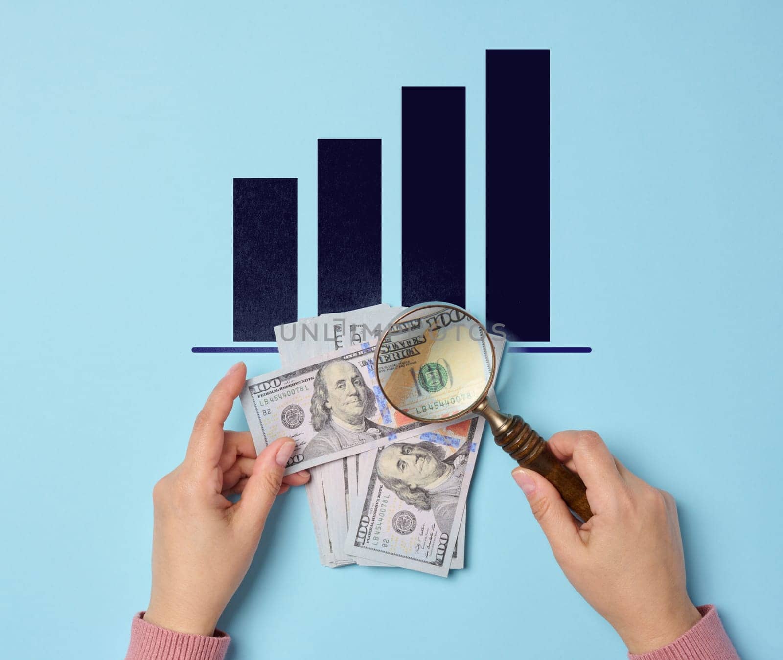 One hundred dollar bills and a woman's hand holding a magnifying glass on a blue background, top view