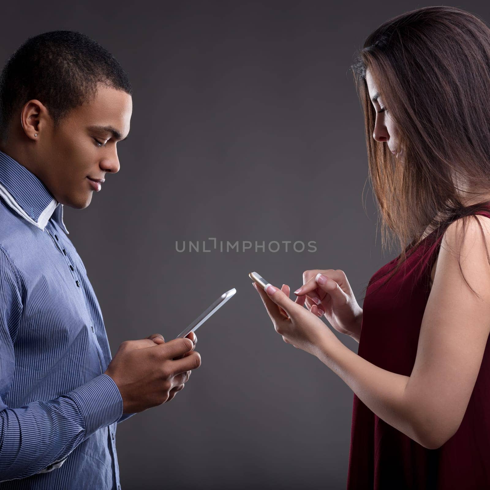 Young couple standing together, absorbed in smartphones, showing the challenge of tech addiction in relationships. Grey background symbolizes disconnect caused by excessive phone use