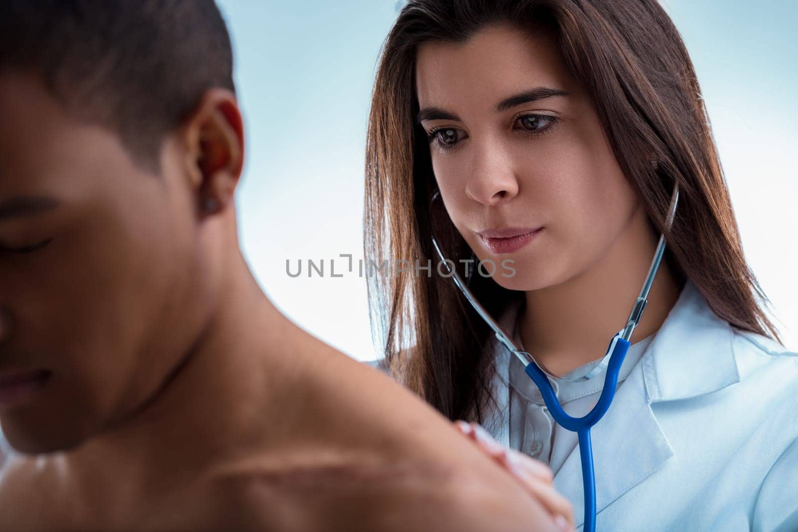 Young doctor is auscultating the lungs of a male patient during a medical examination. The doctor is wearing a white coat and has a stethoscope around her neck