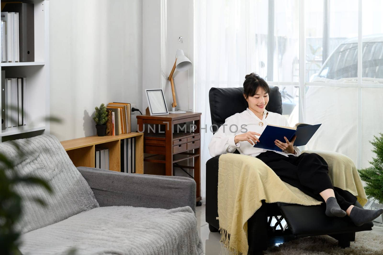 Pretty young woman sitting in a cozy armchair and reading a book with a smile.