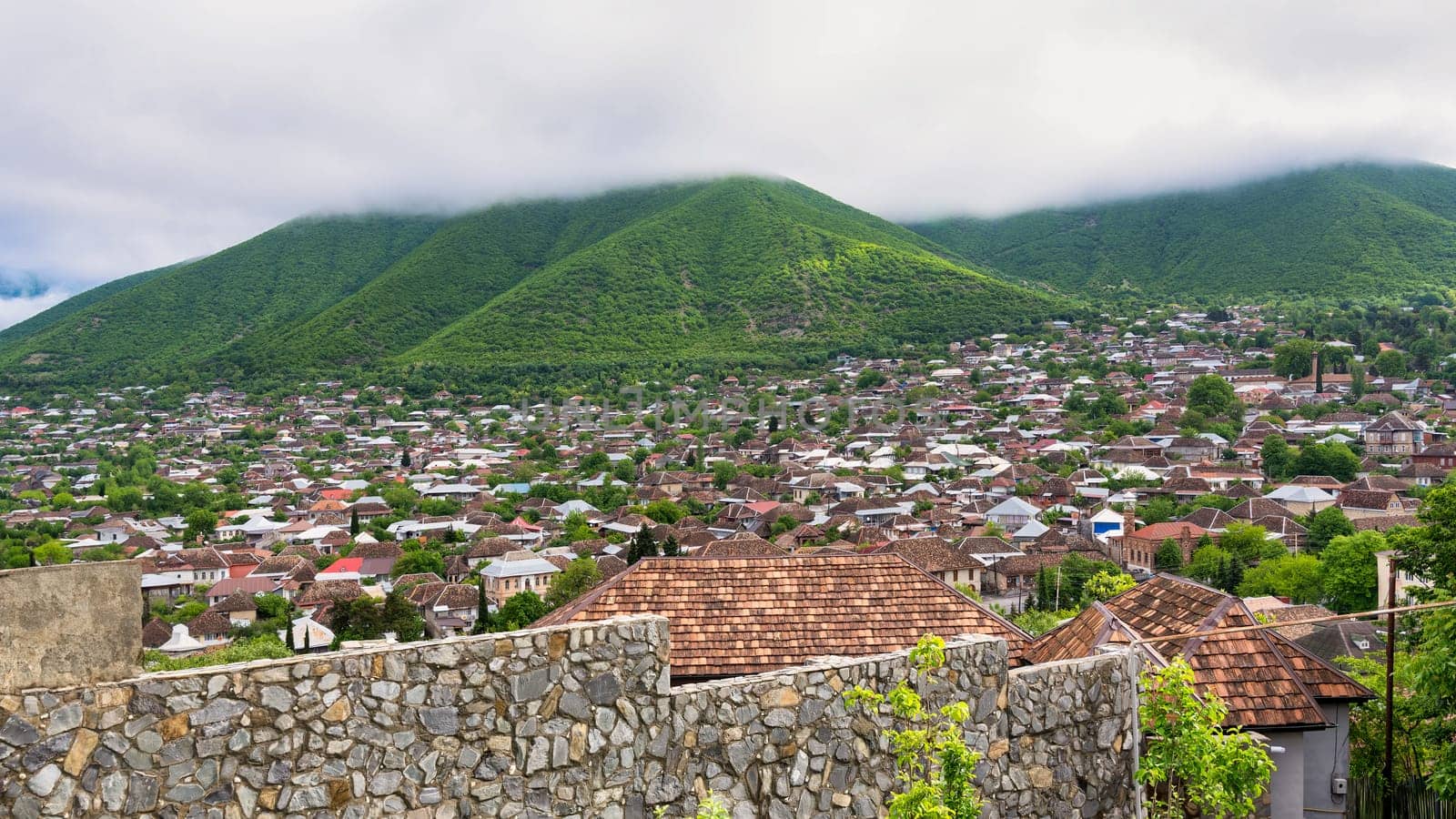 A view of the city of Shaki in Azerbaijan, overlooking a densely populated area with traditional houses and a green mountain in the background. The mountain is covered in fog