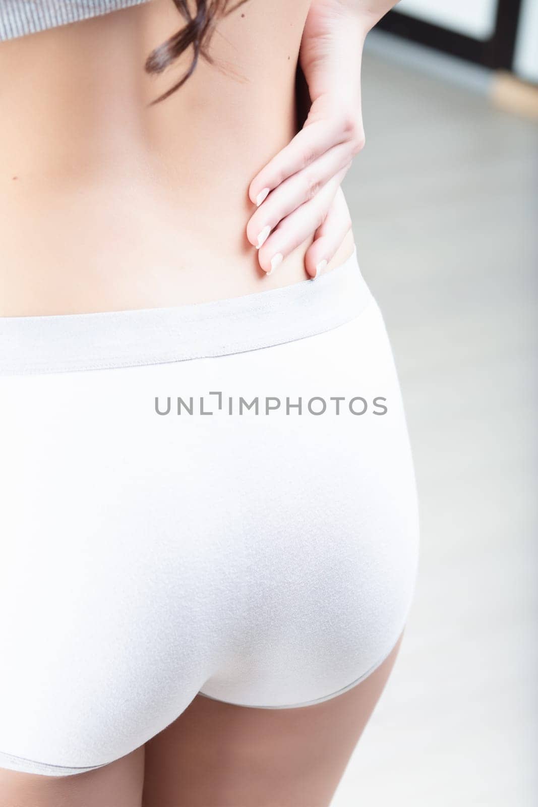 woman in white shorts and a grey top is holding her lower back, suggesting back pain or fatigue. Her hands are positioned to provide support and indicate discomfort in the lumbar area