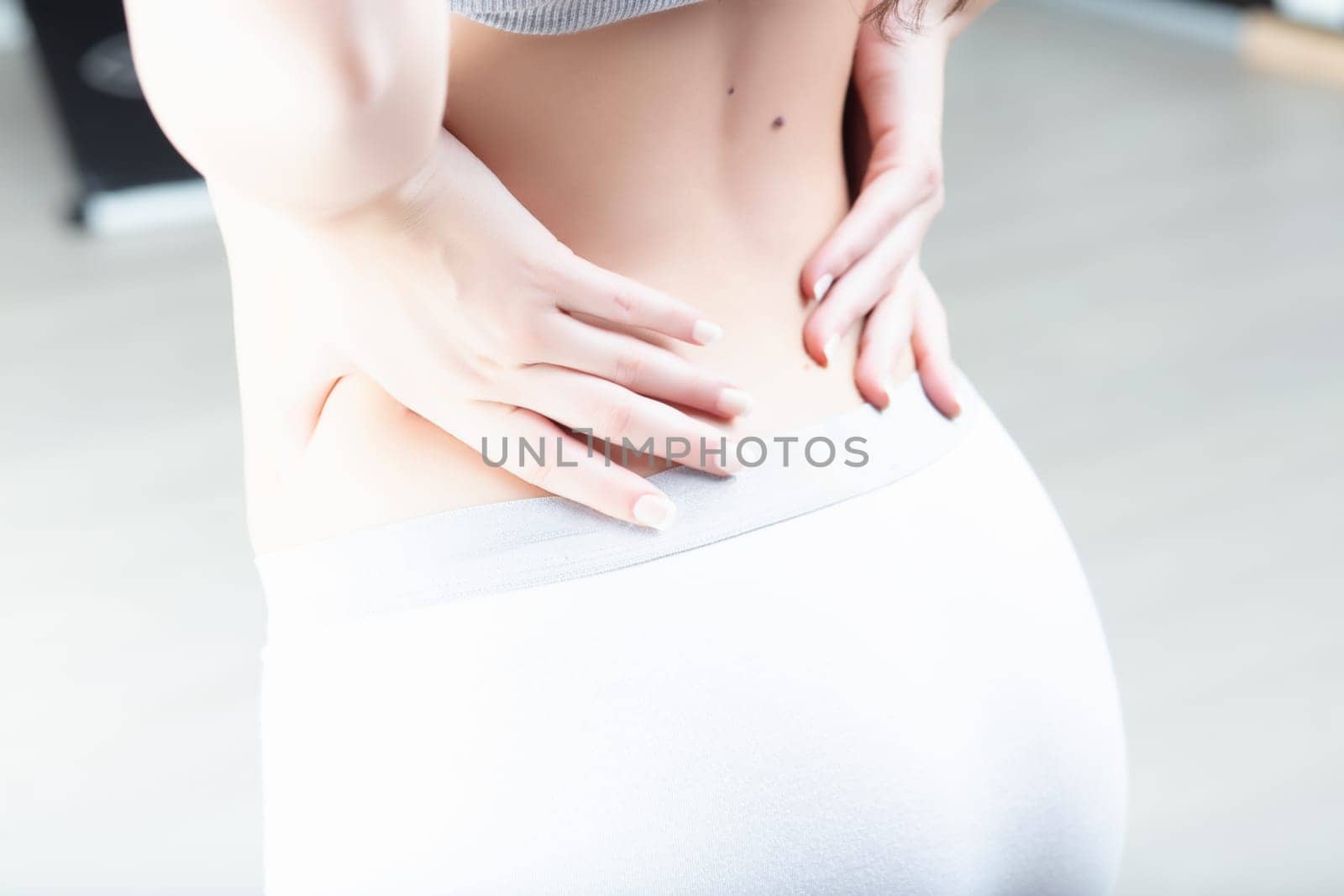 Woman's hands placed on her lower back, wearing casual white shorts and a grey top. This posture suggests she is experiencing discomfort or fatigue in the lumbar region