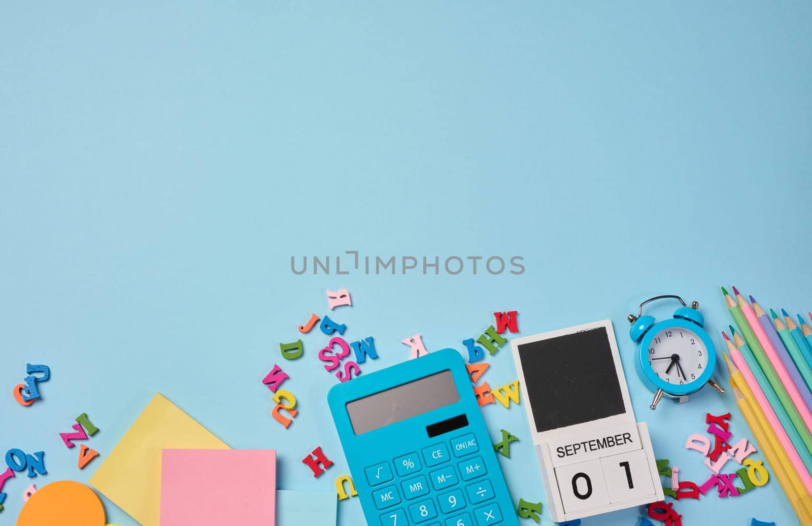 Stationery laid out on a blue background. Multi-colored pencils, calculator, alarm clock, sticky notes and wooden letters, preparing for study or work, organizing workspace