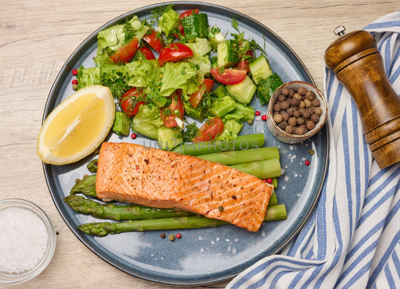 Healthy lunch with grilled salmon with asparagus and fresh tomato and cucumber salad, a row of lemon slice. Food on a blue round plate, top view