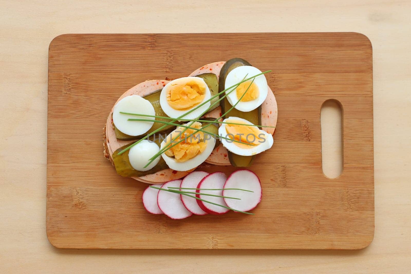 A loaf of bread with sausage, egg, cucumber, radish and chives on a wooden board