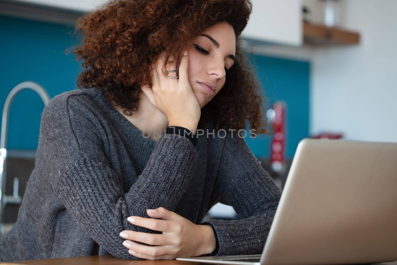 Woman with curly hair rests her face on her hand, staring at her laptop in frustration. The modern kitchen setting emphasizes her impatience, likely caused by buffering delays