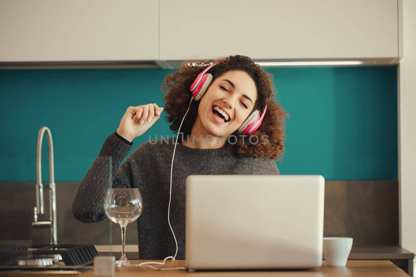 Happy young woman with curly hair is wearing headphones and listening to the music while working on a laptop in the kitchen