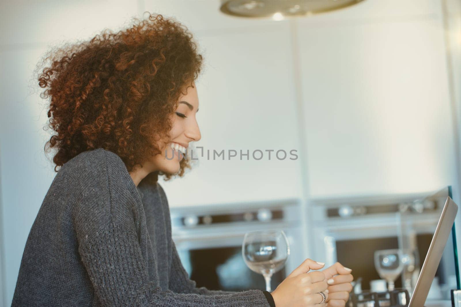 Young woman with curly hair sits at her cozy kitchen counter, smiling and using a laptop, happy and relaxed in her modern apartment, browsing online and connecting with others