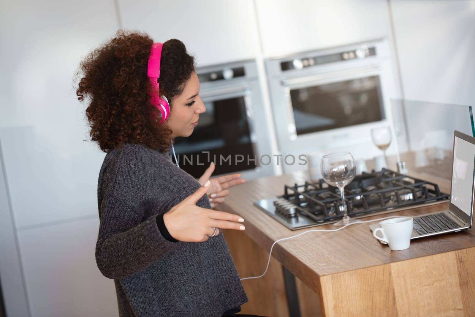 Woman with curly hair wears pink headphones in a modern kitchen, mimicking DJing motions. She sits by a laptop, with a cup and wine glasses nearby, creating a vibrant, club-like atmosphere