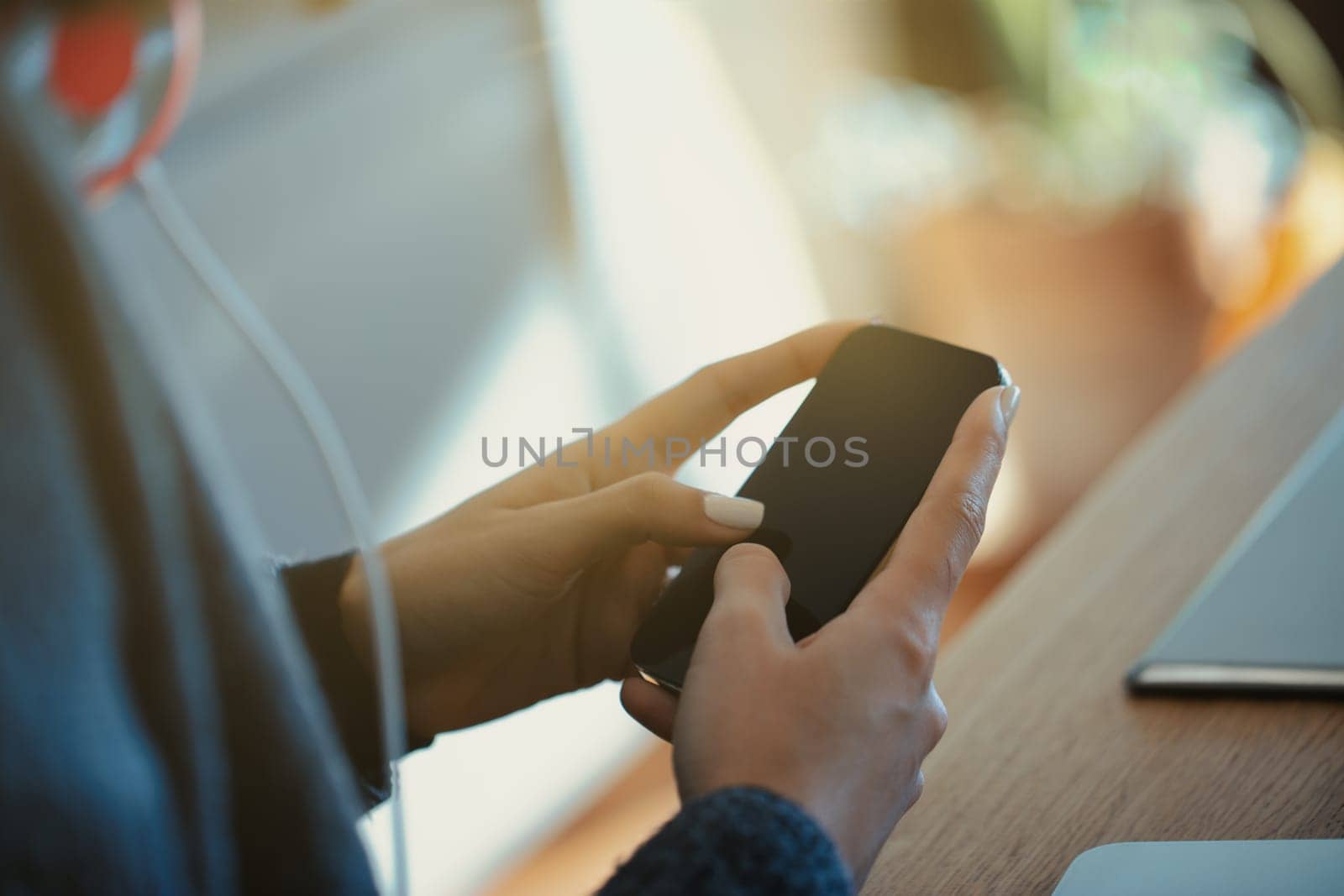 Young woman is holding a smartphone with a blank screen, using her thumb to navigate through the content. The warm light and blurred background create a cozy and inviting atmosphere