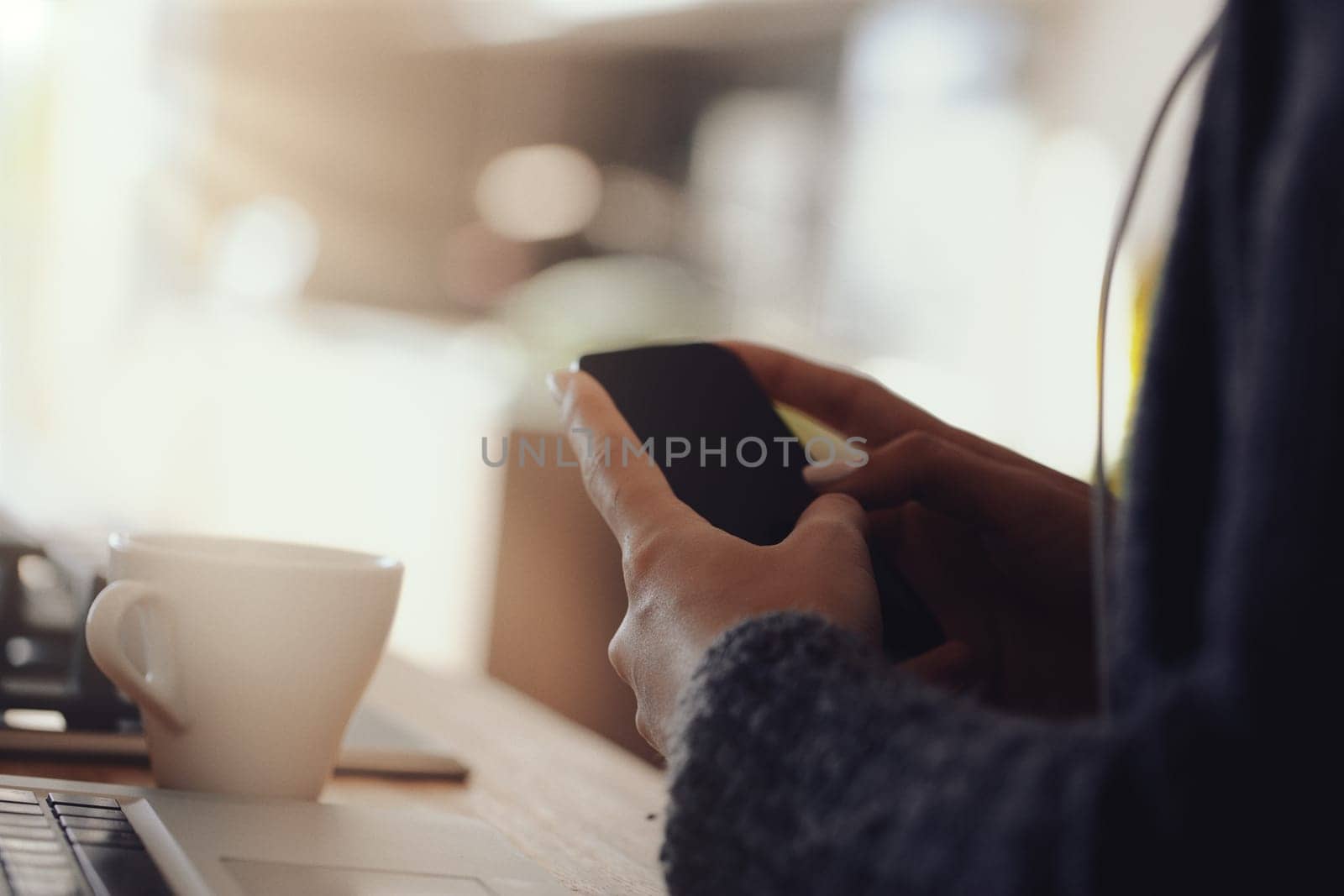 Hands hold a smartphone with a blank screen in a well-lit room. Nearby, a white coffee cup and other objects on a wooden table suggest a calm, casual setting with modern technology