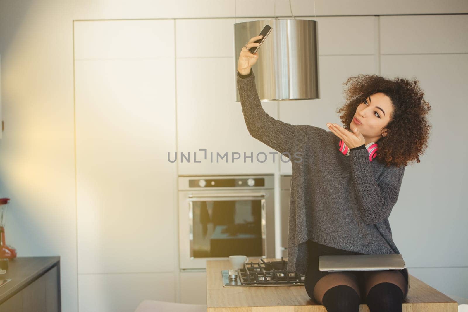 Young woman with curly hair takes a selfie in a bright kitchen, exuding a cheerful vibe. She sends a kiss to the camera, fully immersed in the digital world