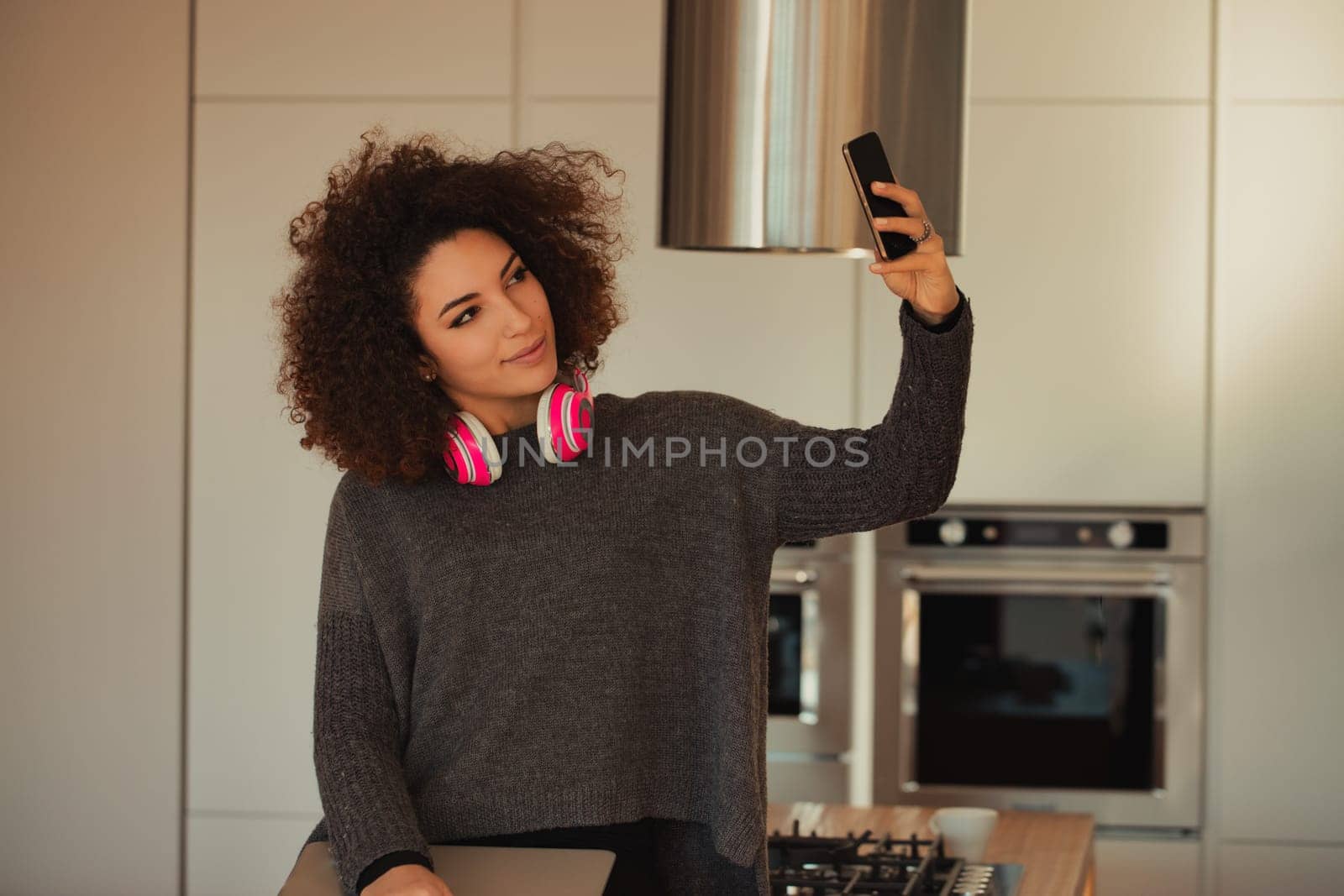 Young woman is taking a selfie in her modern kitchen while wearing headphones, suggesting a fun and carefree atmosphere