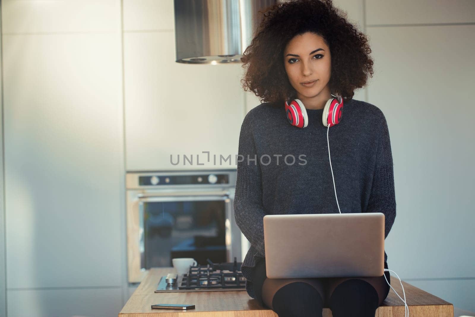 Young woman is working remotely, sitting on her kitchen counter with her laptop. She is wearing headphones and is concentrating on her work