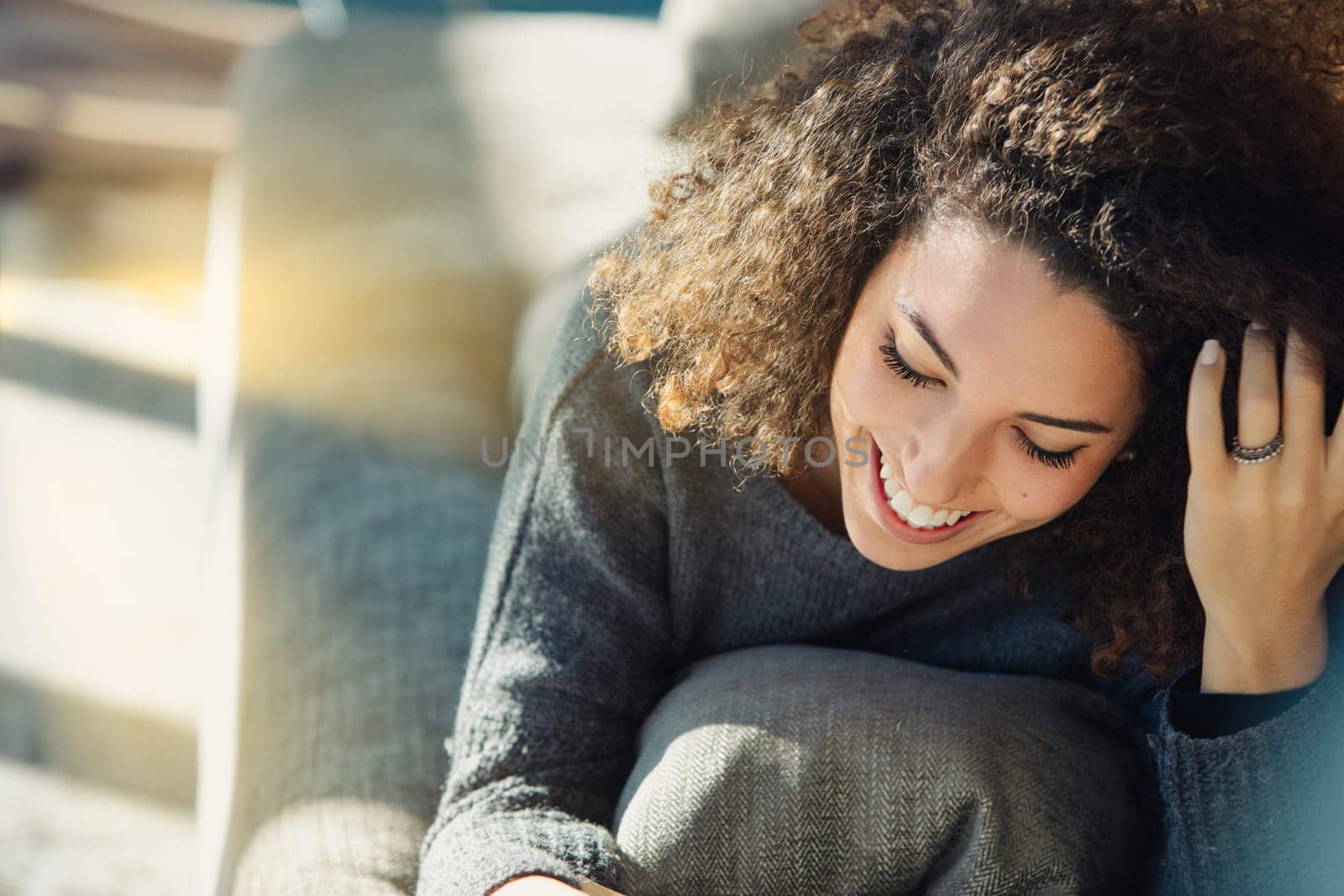 Young woman with curly hair sits happily on a cozy sofa, enjoying a peaceful moment at home. Sunlight adds tranquility, showcasing her joy and positivity in a natural setting