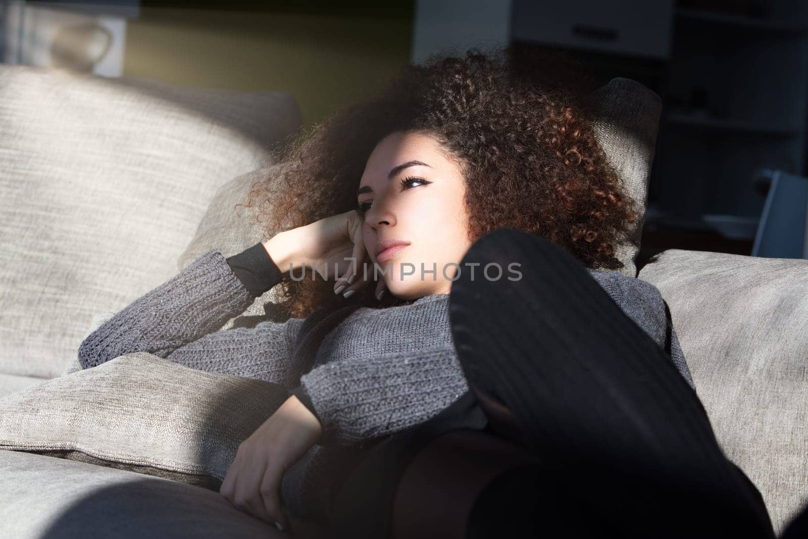 Curly-haired woman lounges on her couch in the living room, lost in thought. Her introspective expression indicates she's reflecting on profound and wonderful ideas, enjoying a moment of quiet contemp