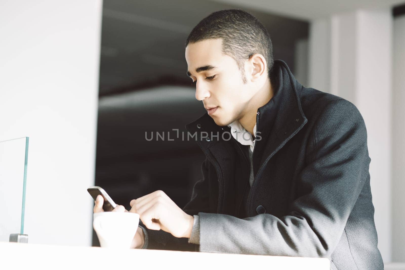 Young man sits at a table in a modern home, using his smartphone during a coffee break, looking focused and serious
