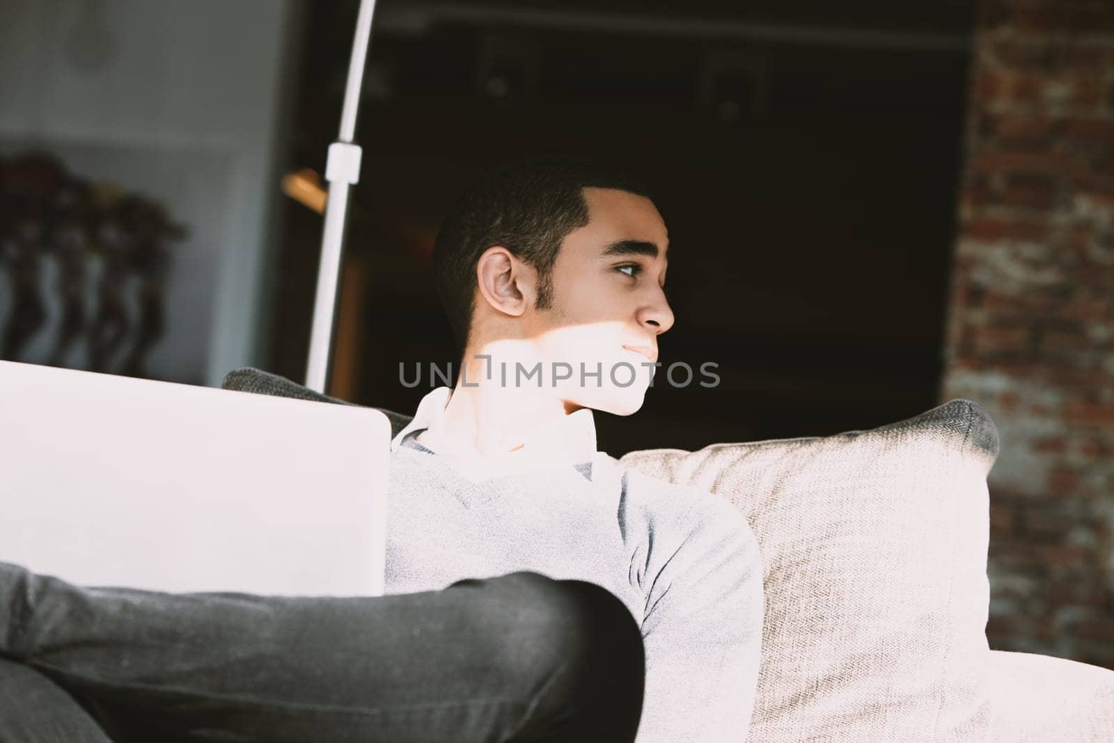 Young man is taking a break from working at home, sitting comfortably on his couch with his laptop resting on his lap