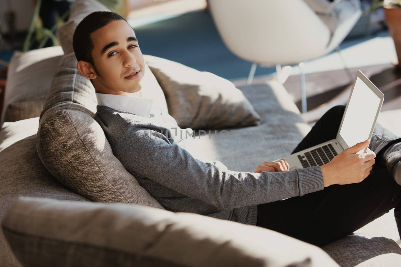 Young professional is working remotely, comfortably seated on a couch in a modern home office, using a laptop with a blank screen