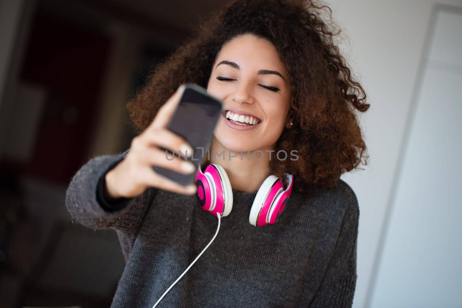Young woman in a cozy kitchen takes a happy selfie with pink headphones, enjoying music and sharing her joy on social media