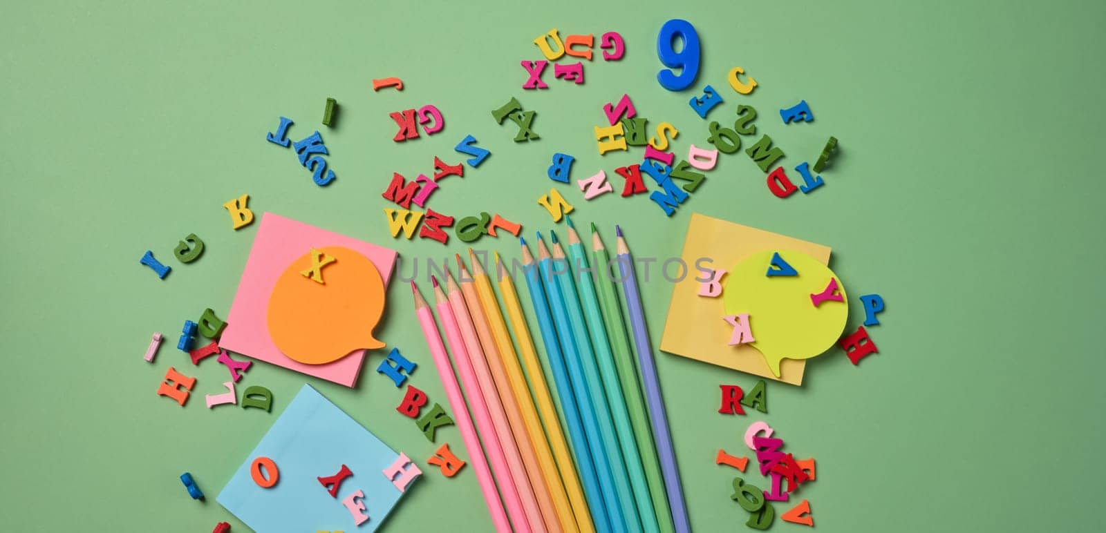 Stationery laid out on a green background. Multi-colored pencils ,  sticky notes and wooden letters, preparing for study or work, organizing workspace