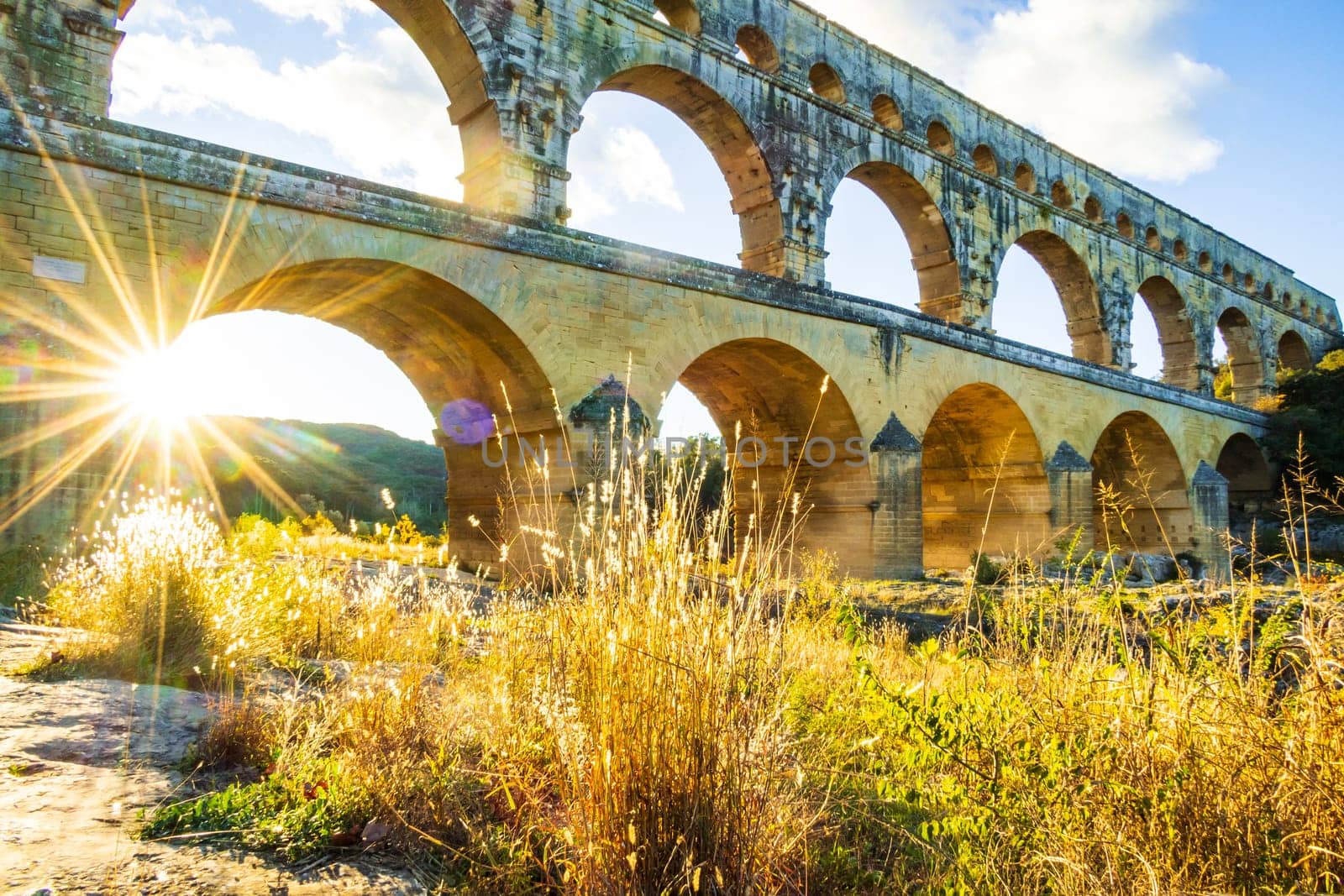 The magnificent Pont du Gard backlit by the setting sun. Ancient Roman three-tiered aqueduct bridge built in the 1st century AD to carry water to the Roman colony of Nemausus (Nîmes). Photograph taken in Vers-Pont-du-Gard, Provence, southern France.