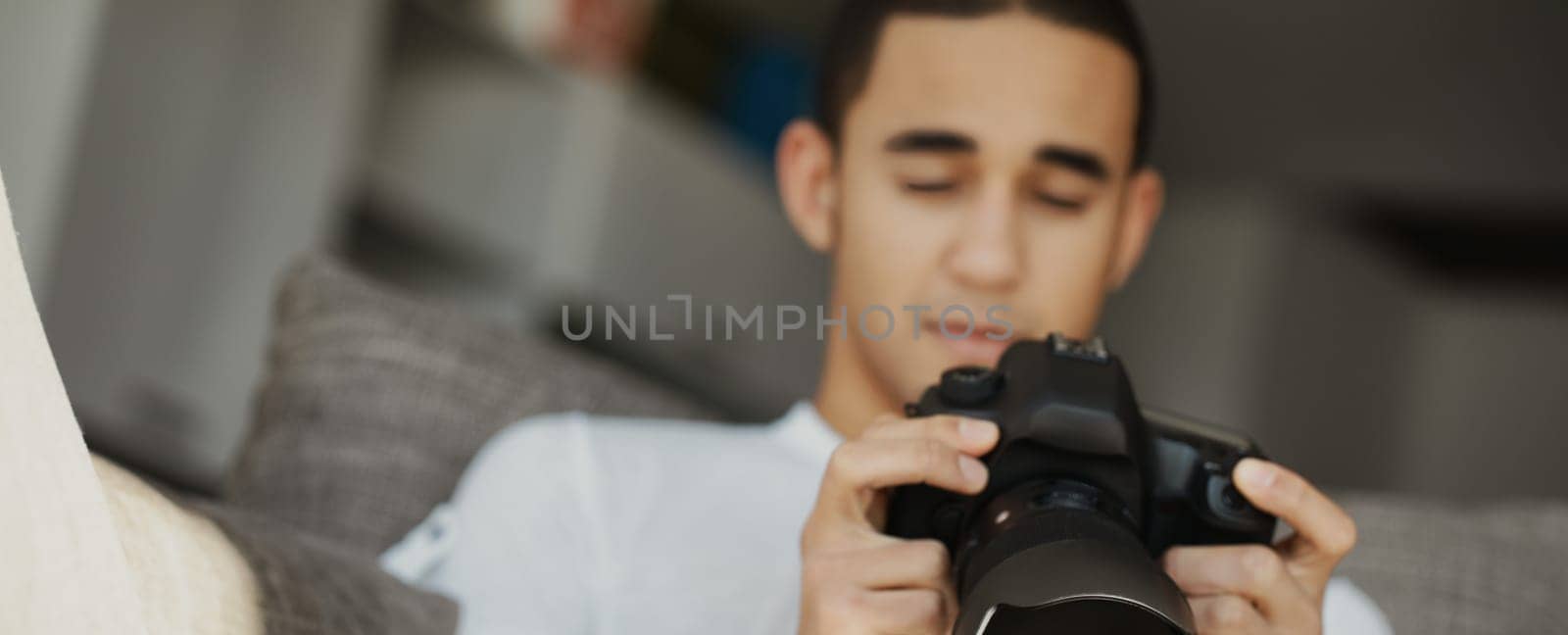 Young man adjusts a professional camera on a sofa at home, immersed in photography. Natural light enhances his creative focus
