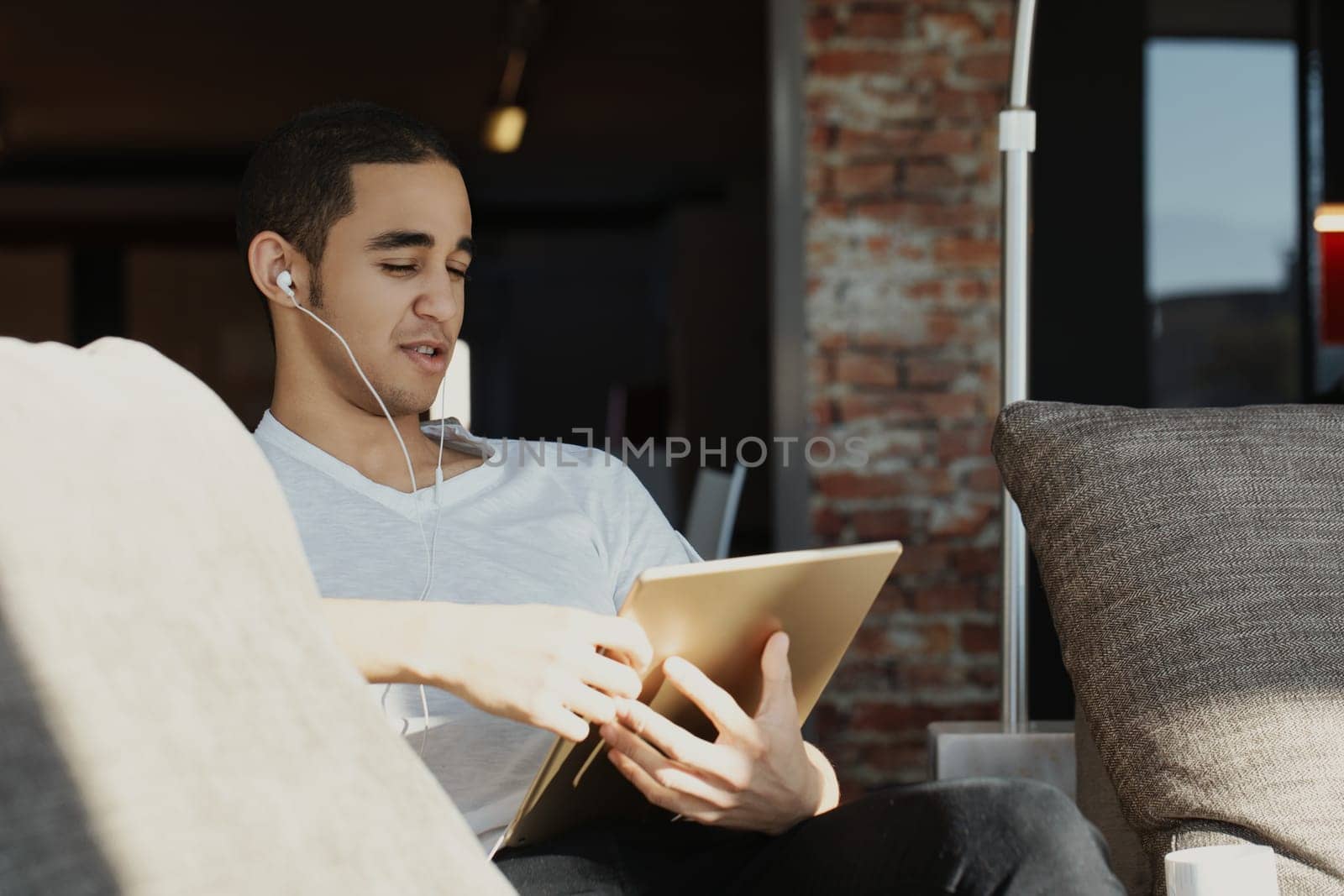 Young man in a white t-shirt, wearing earphones, looks skeptical while using a tablet on a sofa. His critical expression shows doubt about what he's seeing online. The brick wall adds a modern touch