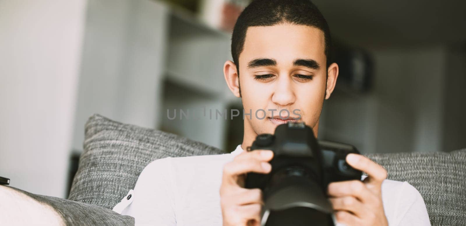 Sitting on a sofa, a young man in a white t-shirt carefully looks at the DSLR display, reviewing his photos. The relaxed setting and soft lighting emphasize his calm demeanor