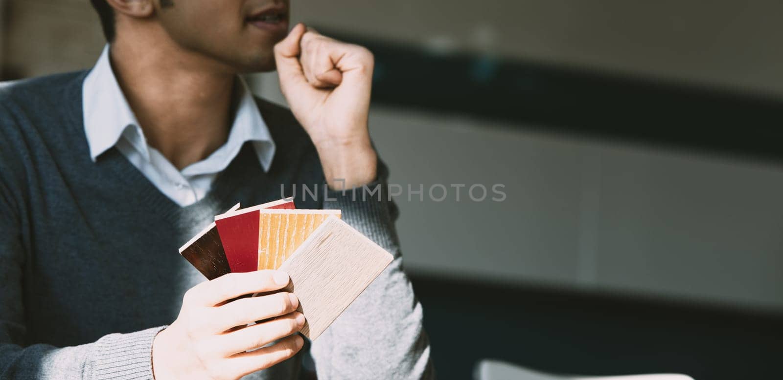 In a sunlit room, an unrecognizable man with short hair and a gray sweater over a white shirt examines color samples. His thoughtful expression is emphasized by the warm, natural light and modern deco