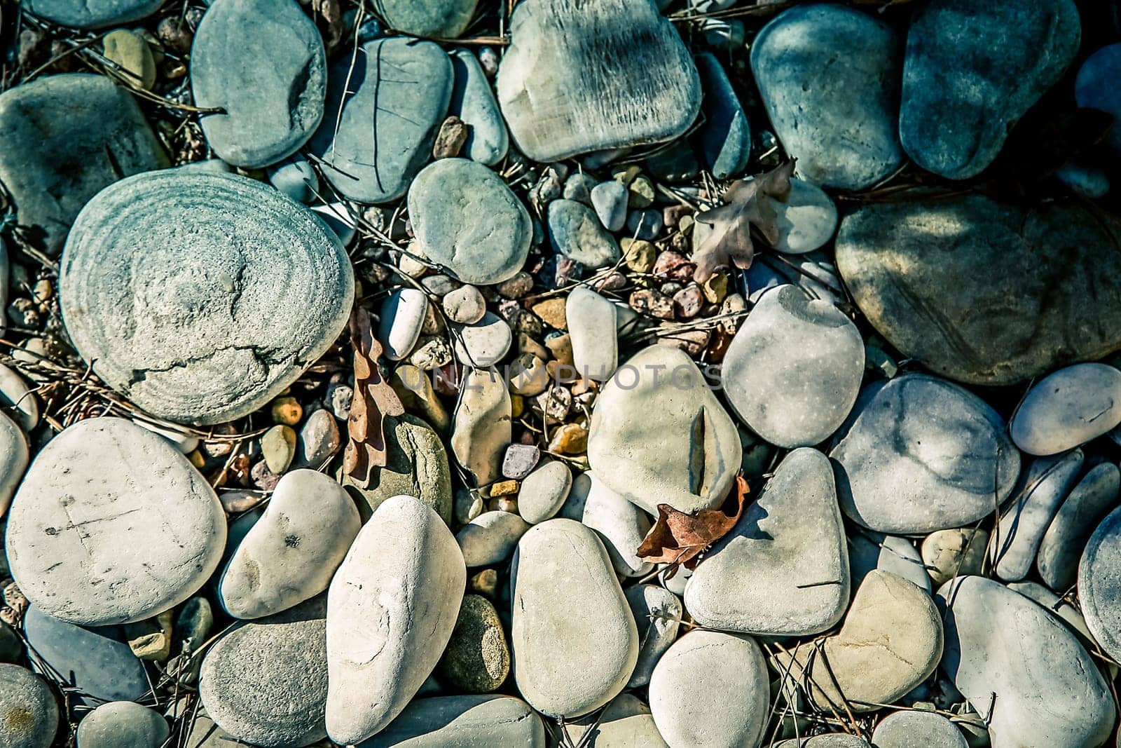 A collection of stones and pebbles of different sizes on the ground with sunlight and shadows, as well as an autumn leaf among them, background image