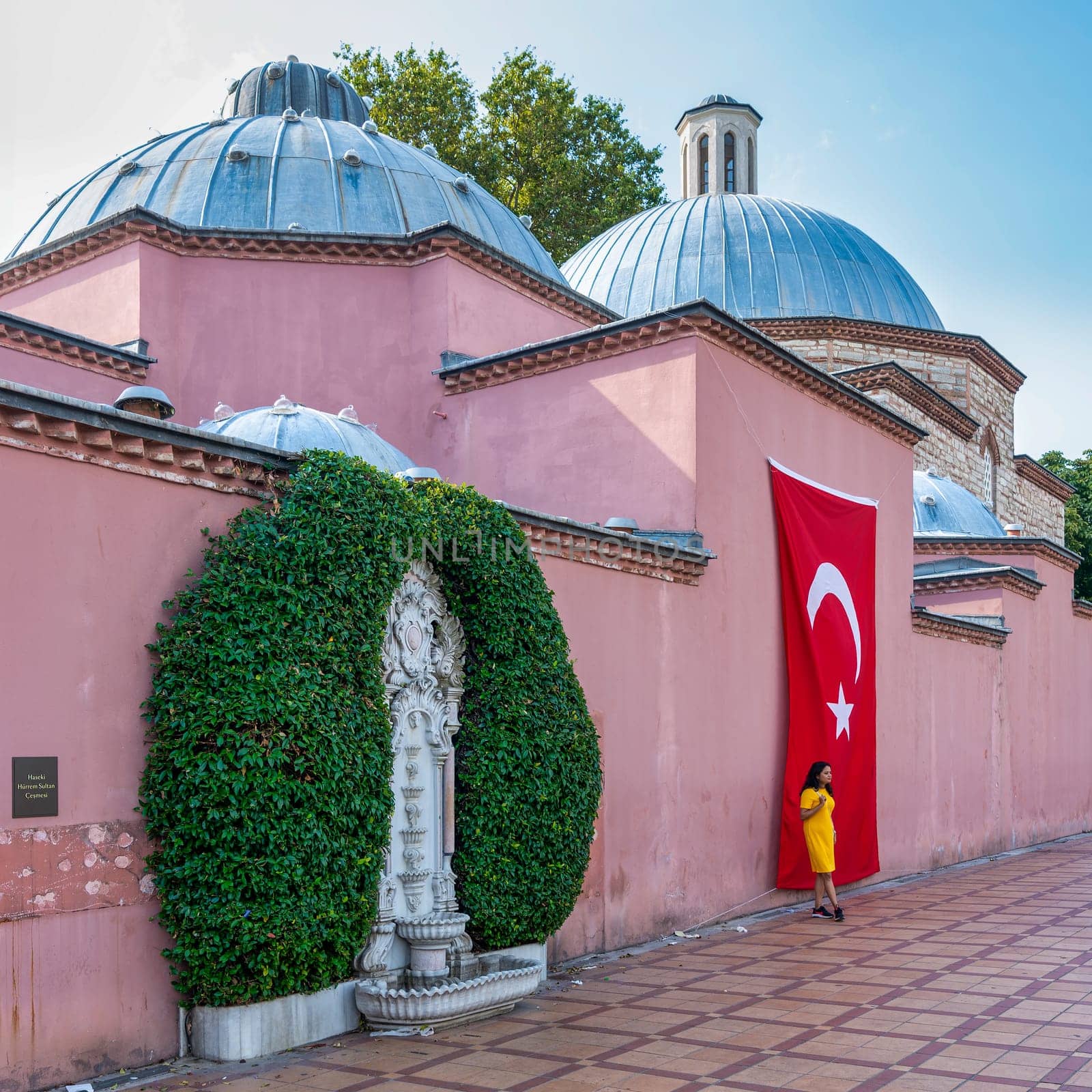 Asian woman in a yellow dress stands beside a Turkish flag hanging on the wall of the Hagia Sophia Hurrem Sultan Bathhouse