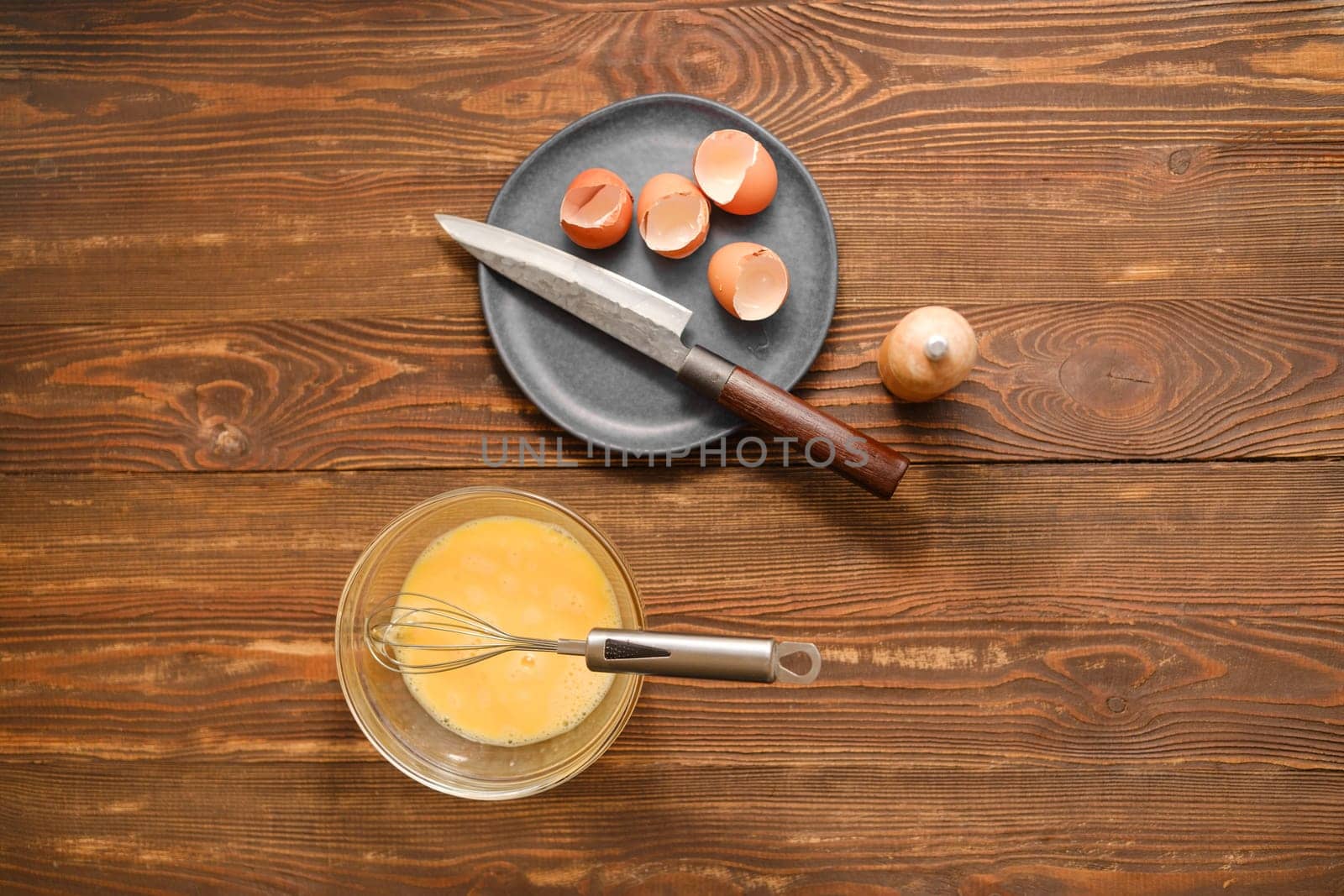 Top view of rustic wooden table with a bowl with beaten eggs and a plate with broken eggshells and a knife