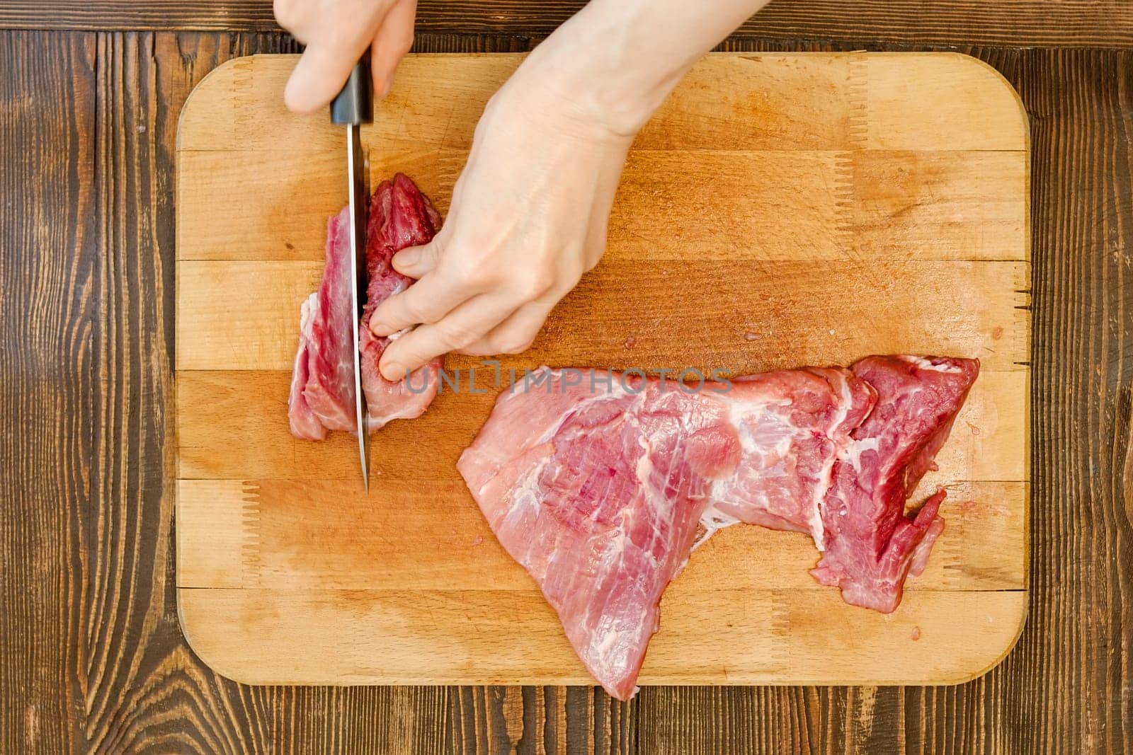 Top view of hands of unrecognizable man slicing pork shoulder on wooden cutting board