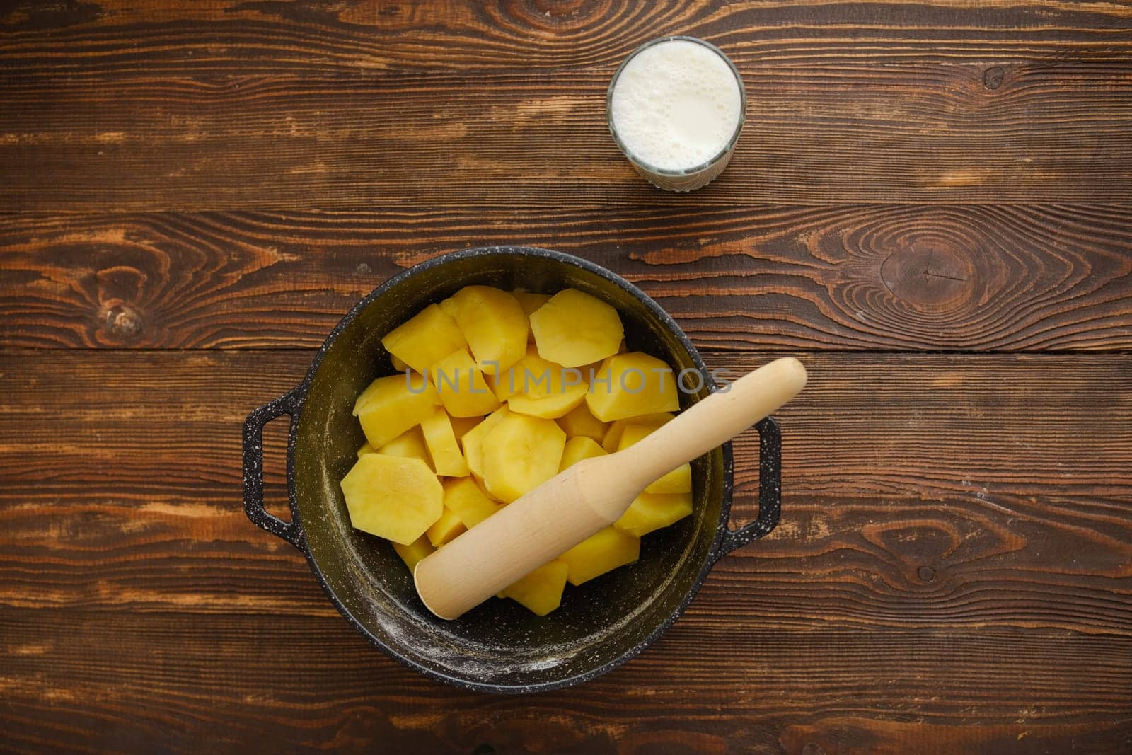 Top view of a kitchen table with boiled potatoes in a pot, potato masher and glass of warm milk
