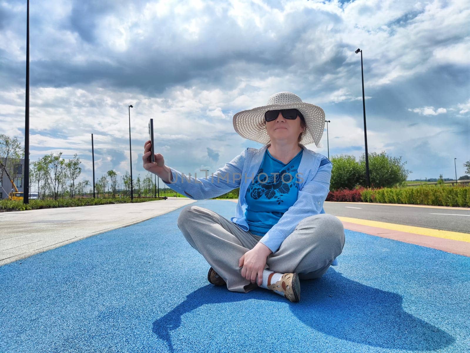 A woman with vitiligo is sitting cross-legged on a colorful pathway, wearing a large-brimmed sun hat and sunglasses while taking a selfie. The sky is partly cloudy.