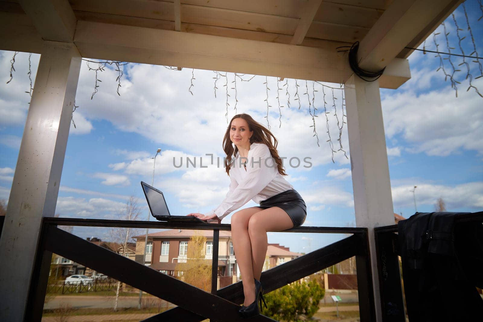 A woman is focused on her laptop while perched on a railing in an outdoor setting on a sunny day.